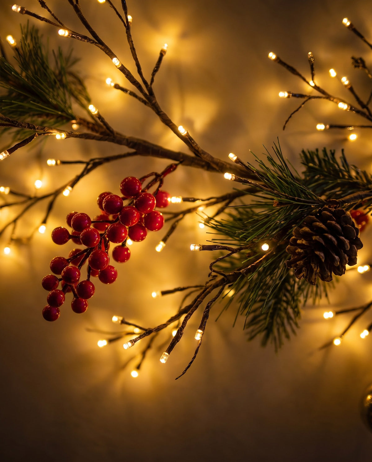 Close-up of red berries, pine needles, a pinecone and tiny warm-white bulbs on the branch