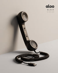 Aloo in black standing against a light grey wall, its coiled USB-C cord on the floor