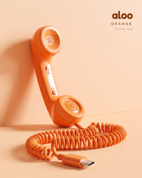 Aloo in orange standing against a peach wall, its coiled USB-C cord on the floor