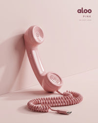 Aloo in pink standing against a blush wall, its coiled USB-C cord on the floor
