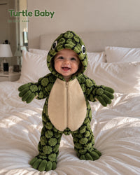 Smiling baby standing on a bed in the green Turtle Baby onesie with a cream tummy and turtle-eye hood